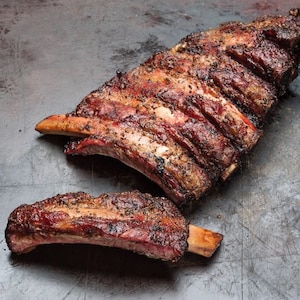 May include: Two portions of cooked beef ribs, with visible bones, are presented on a dark, textured surface. The ribs have a dark, seasoned crust with visible pepper and a reddish-brown interior, suggesting they are freshly cooked.