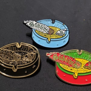 May include: Three enamel pins featuring a blue, black, and red ashtray with a joint inside. Each pin has the word "Sublime" written on the joint. The pins are on a dark gray surface.