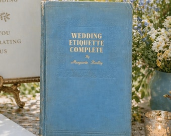 May include: A vintage, light blue book titled "Wedding Etiquette Complete" by Marguerite Bentley. The book is displayed on a table with a white notepad, pen, and a lit candle in a glass holder. Floral arrangements and a welcome sign are in the background.