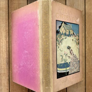 May include: A vintage hardcover book titled All in a Garden Fair by Alice T.A. Quackenbush. The cover is light pink with a central rectangular illustration depicting a cottage, a person tending to rose bushes, and the title in black lettering.