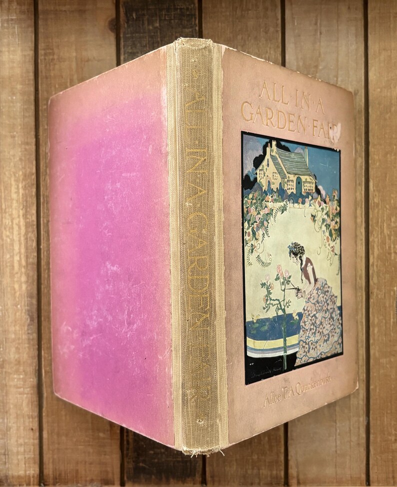 May include: A vintage hardcover book titled All in a Garden Fair by Alice T.A. Quackenbush. The cover is light pink with a central rectangular illustration depicting a cottage, a person tending to rose bushes, and the title in black lettering.