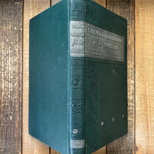 May include: A vintage, dark teal book titled "Chemical Gardening for the Amateur" by Connors and Tiedjens. The cover features embossed floral designs and the publisher's name, Wise & Co. The book is set against a wooden background.