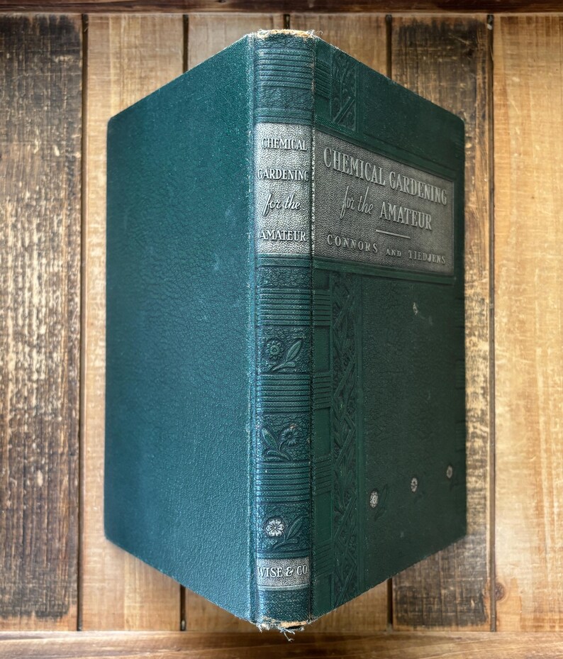 May include: A vintage, dark teal book titled "Chemical Gardening for the Amateur" by Connors and Tiedjens. The cover features embossed floral designs and the publisher's name, Wise & Co. The book is set against a wooden background.