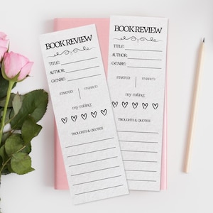 May include: Two white book review cards with black text and heart-shaped rating symbols. The cards have sections for title, author, genre, and thoughts. A pink notebook and a pencil are also in the image.