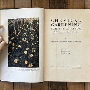 May include: An open book with the title "CHEMICAL GARDENING FOR THE AMATEUR" on the right page. The left page features a color photograph of a greenhouse filled with flowering plants. The book is on a wooden surface.