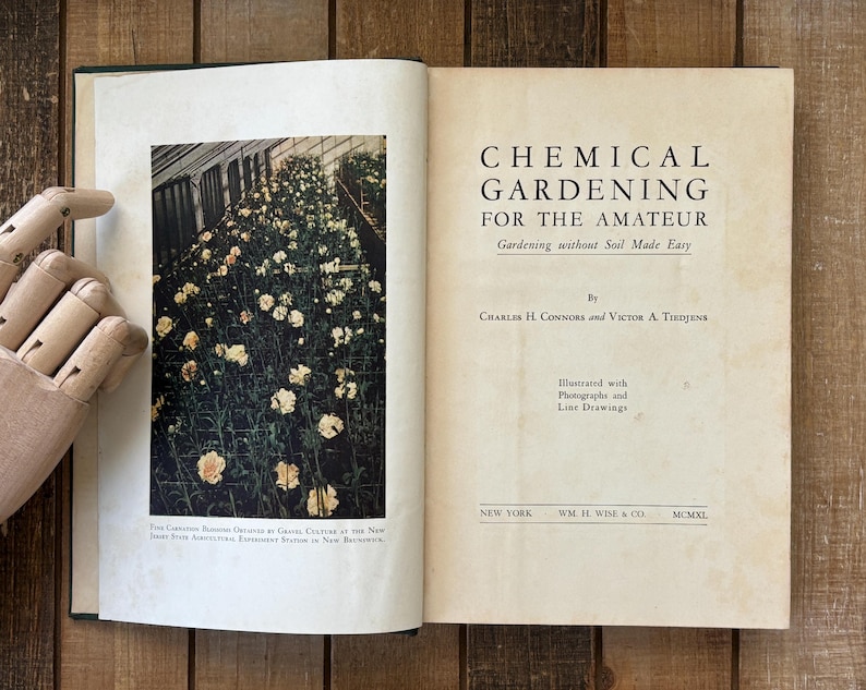 May include: An open book with the title "CHEMICAL GARDENING FOR THE AMATEUR" on the right page. The left page features a color photograph of a greenhouse filled with flowering plants. The book is on a wooden surface.