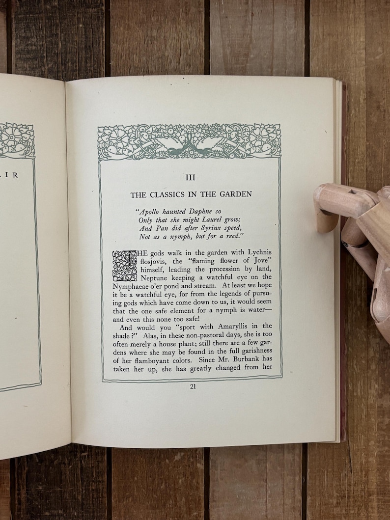 May include: An open book page titled III THE CLASSICS IN THE GARDEN with a decorative green border and a large drop cap letter T. The text discusses Apollo, Daphne, Pan, and Syrinx, with the page number 21 at the bottom center of the page.