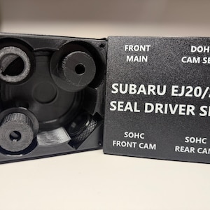 Op de afbeelding: Een zwarte Subaru EJ20/25 Seal Driver Set. De set bevat verschillende zwarte componenten, waaronder cilindrische stukken en een rechthoekige doos met de tekst "FRONT MAIN", "DOHC CAM SEAL", "SOHC FRONT CAM" en "SOHC REAR CAM".