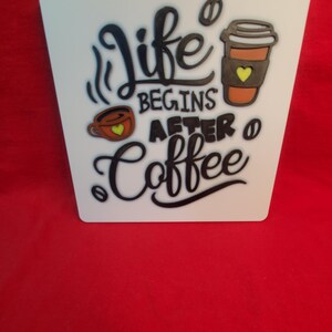 May include: White, square-shaped sign with the phrase "Life Begins After Coffee" in black script. The sign features coffee cup illustrations in brown and orange with yellow hearts. A small hole is at the top for hanging.