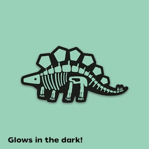 K&ouml;nnte beinhalten: Schwarze Silhouette eines Stegosaurus-Skeletts mit einer Leucht-im-Dunkeln-Funktion. Der Dinosaurier hat einen stacheligen R&uuml;cken und einen langen Schwanz. Der Text "Glows in the dark!" befindet sich unter dem Bild.
