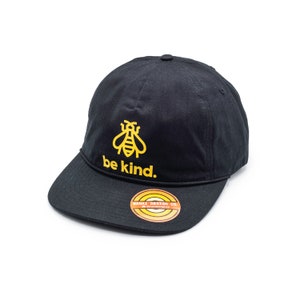 Op de afbeelding: Zwarte baseballpet met een gele bij en de tekst "be kind." De pet heeft een geel en oranje label met de tekst "MANAZ DESIGN CO."