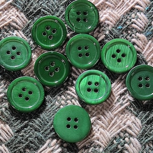 May include: A collection of ten round, glossy green buttons with four holes each. The buttons are arranged on a textured, patterned fabric in shades of green, brown, and beige. The buttons vary slightly in shade.