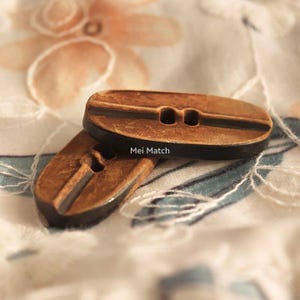 May include: Two oval-shaped buttons with a brown, wood-like textured finish and a dark edge. Each button features a central horizontal groove and two holes for attachment. The text Mei Match is printed in white on the top button surface.