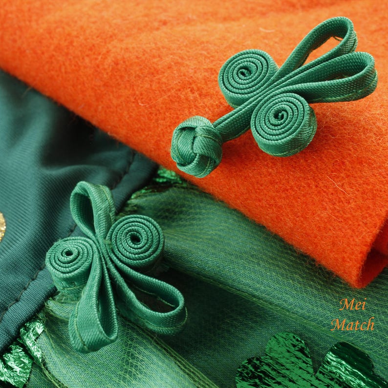 May include: Two green fabric loops with a knotted center, resembling a shamrock, are attached to a green and orange fabric. The fabric is folded and the loops are on the top of the fold. The text "Mei Match" is visible in the bottom right corner of the image.