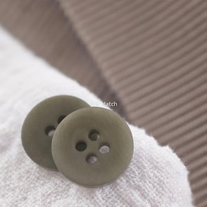 May include: Two round, olive-green buttons with four holes in the center, featuring the text Mei Match printed in white. The buttons have a smooth, matte finish and are positioned on a white, textured fabric surface with a brown ribbed fabric nearby.