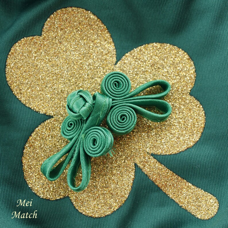May include: A green fabric knot with a gold glitter shamrock design on a green background. The knot is a traditional Chinese knot, also known as a "lucky knot".