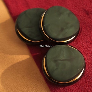 Puede incluir: Tres botones circulares con una superficie verde oscuro mate y una sutil textura marmolada. Cada botón está acentuado con una fina banda metálica de color dorado pulido en el borde. El texto Mei Match es visible entre los botones.