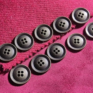May include: A collection of ten round, four-hole buttons with a metallic bronze finish arranged in two diagonal rows on a textured, deep magenta fabric surface. Each button features a slightly recessed centre and a smooth, raised outer rim.