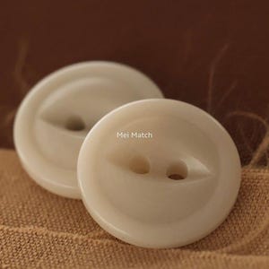 May include: Two round, off-white buttons with a smooth, glossy finish. Each button features two central holes recessed within a shallow, eye-shaped indentation. The text "Mei Match" is printed in small, dark letters on the surface of the front button.