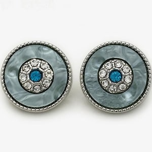 May include: A pair of round earrings with a silver-colored metal border. The earrings have a light blue marbled center with a circle of clear crystals surrounding a blue center stone. The earrings are set against a white background.