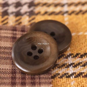 May include: Two round, dark brown buttons with a smooth, polished finish and four central holes. The text Mei Match is printed in white on the surface of the front button. The buttons have a slightly concave centre and a raised outer rim.