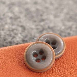 Peut inclure: Deux boutons ronds avec un centre gris marbré lisse et un bord brun clair. Chaque bouton comporte quatre trous au centre pour la fixation. Le texte Mei Match est imprimé en blanc entre les deux boutons sur la surface orange texturée.