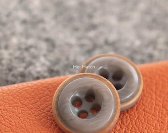 Op de afbeelding: Twee ronde knopen met een glad, grijs, gemarmerd midden en een lichtbruine rand. Elke knoop heeft vier gaten in het midden voor bevestiging. De tekst Mei Match is in het wit gedrukt tussen de twee knopen op het oranje, getextureerde oppervlak.