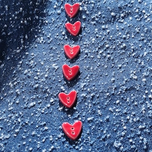 Op de afbeelding: Een verticale rij van zes kleine, glanzende rode hartvormige knopen, elk met twee centrale gaten voor bevestiging, gerangschikt in een rechte lijn tegen een achtergrond van gestructureerde, donkerblauwe spijkerstof met witte spikkels.