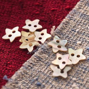 May include: A collection of star-shaped buttons crafted from iridescent mother-of-pearl material. Each button features two central holes for attachment and displays a smooth, glossy finish with subtle colour variations. The text Mei Match is visible.