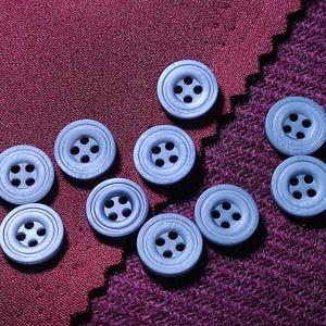 May include: A collection of nine round, light blue buttons, each featuring four central holes and a double-ringed border design. The buttons are scattered across a surface that transitions from a smooth, dark red fabric to a textured purple material.