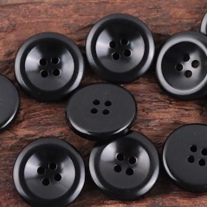 May include: A collection of glossy black buttons with four holes each. The buttons are round and have a slightly concave center. The image shows a variety of sizes and arrangements of the buttons, suitable for sewing or crafts.