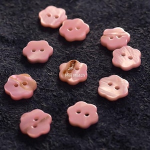 Op de afbeelding: Een verzameling roze, bloemvormige knopen met elk twee gaatjes. De knopen hebben een parelmoerglans en liggen op een donkere, getextureerde ondergrond. De tekst "Mei Match" is zichtbaar op één knoop.