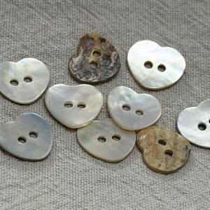 Pode incluir: Uma coleção de botões em forma de coração com vários acabamentos, incluindo madrepérola branca iridescente e material castanho texturizado semelhante a madeira. Cada botão tem dois orifícios centrais para fixação, dispostos de forma dispersa.