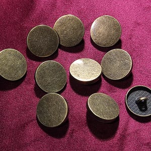 Puede incluir: Colección de botones redondos de latón antiguo con superficie texturizada. Los botones están dispersos sobre una tela rojo oscuro, mostrando su apariencia envejecida y brillo metálico. Un botón muestra la parte trasera con un vástago negro.