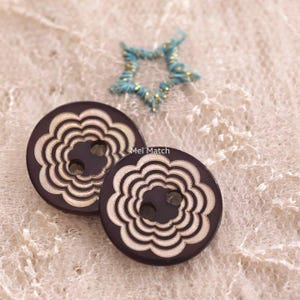 Peut inclure: Deux boutons ronds violet foncé avec un motif floral crème aux bords festonnés concentriques et deux trous centraux. Le texte Mei Match est visible entre les boutons. Un petit accent en forme d'étoile bleu et or est au-dessus.