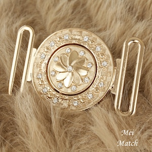 Puede incluir: Hebilla de cinturón de metal dorado con un diseño redondo y adornado que presenta una flor con brillantes transparentes. La hebilla tiene dos lazos para sujetar a un cinturón.
