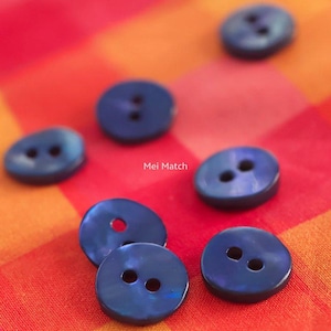Puede incluir: Un conjunto de siete botones redondos de color azul oscuro, cada uno con dos agujeros centrales. Los botones tienen una superficie lisa y ligeramente reflectante y están dispersos por el encuadre. El texto Mei Match es visible en el centro.