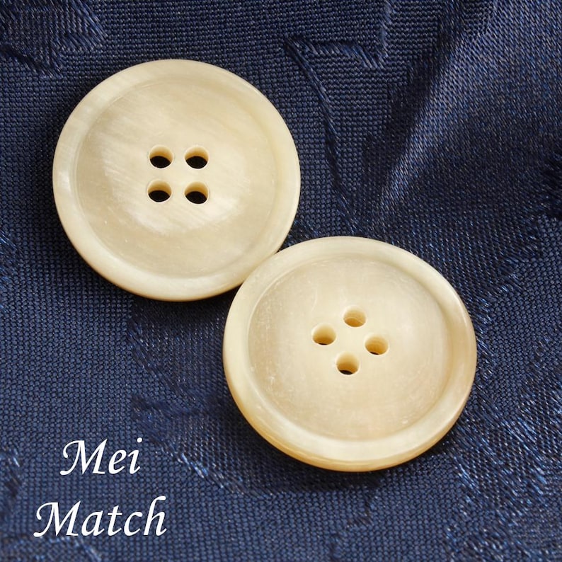 May include: Two off-white, round buttons with four holes each, set against a textured navy blue fabric. The buttons have a subtle, pearlescent sheen. The text "Mei Match" is visible at the bottom of the image.