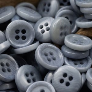 Op de afbeelding: Een close-up van een stapel lichtblauwe plastic knoppen, elk met vier gaten. De knoppen zijn glad en hebben een licht getextureerd oppervlak.
