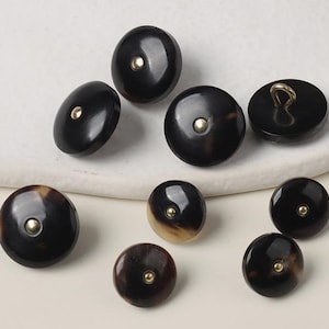 May include: A selection of dark brown buttons with a glossy finish. Each button has a small, gold-coloured centre. Some buttons have a lighter, mottled pattern. The buttons vary in size and shape, with some having a loop on the back.