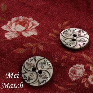 May include: Two white mother of pearl buttons with a floral design and two holes. The buttons are on a red fabric with a floral pattern.
