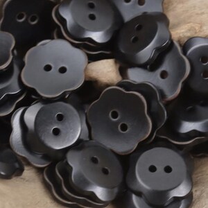 Puede incluir: Un conjunto de botones negros con forma de flor, cada uno con dos agujeros. Los botones tienen un centro ligeramente elevado y un borde festoneado. Fabricados con un material liso y mate.