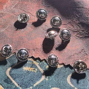 Könnte beinhalten: Eine Ansammlung von neun runden, silberfarbenen Knöpfen, die jeweils eine glatte, reflektierende Kuppel in der Mitte und einen strukturierten, seilartigen Rand aufweisen. Diese metallischen Verschlüsse sind verstreut angeordnet und zeigen ihre polierte Oberfläche.