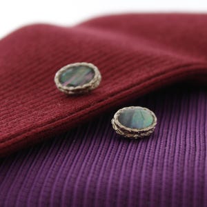 Könnte beinhalten: Zwei ovale Knöpfe mit einem silbernen Metallrahmen und einem dunkel schillernden Kern. Die Knöpfe befinden sich auf einem burgunderfarbenen und violetten Stoff.