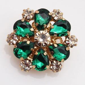Puede incluir: Un adorno dorado en forma de flor con piedras de color verde esmeralda y cristales transparentes. La piedra central es transparente, rodeada de ocho piedras verdes en forma de lágrima. Piedras más pequeñas y transparentes se colocan alrededor del perímetro.