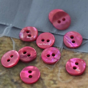 Può includere: Una collezione di dieci bottoni rotondi a due fori in un vivace colore rosa magenta con una finitura lucida e iridescente. Ogni bottone presenta una superficie liscia e leggermente bombata. Il testo Mei Match è visibile al centro del gruppo.