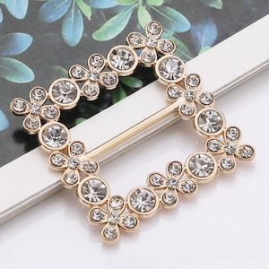 May include: Gold-toned rectangular buckle adorned with clear, sparkling rhinestones. The buckle features a floral design with larger round rhinestones at intervals. The overall design is symmetrical and elegant, suitable for various fashion accessories.