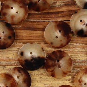 May include: A collection of round buttons with two holes each. The buttons have a marbled appearance with shades of brown and tan. They are arranged on a wooden surface, showcasing their texture and color variations.