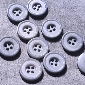 Puede incluir: Un conjunto de diez botones redondos de color gris claro, cada uno con un borde exterior liso y ligeramente elevado y cuatro agujeros centrados para su fijación. Los botones tienen un acabado mate uniforme y están dispuestos dispersos.