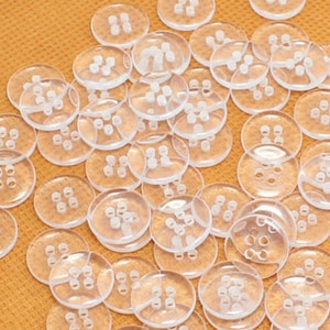May include: A large group of clear plastic buttons with four holes each. The buttons are scattered on a light orange surface.